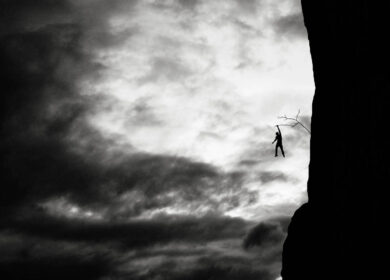 Defying the danger of life Man holding onto branch sticking out of rock wall