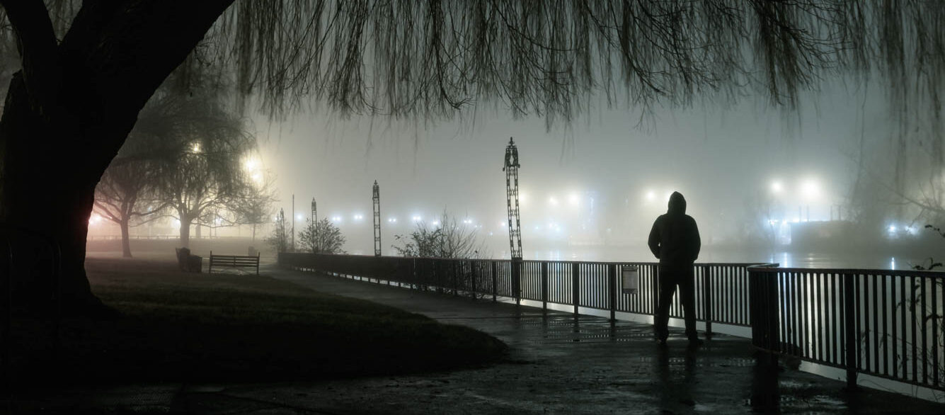 A mysterious moody hooded figure silhouetted against street lights by a river on a foggy atmospheric winters night A mysterious moody hooded figure silhouetted against street lights by a river on a foggy atmospheric winters night