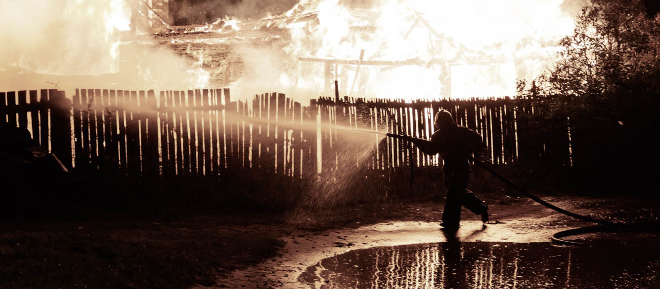 Firefighter Spraying Water From Hose On Burning House At Night Firefighter Spraying Water From Hose On Burning House At Night