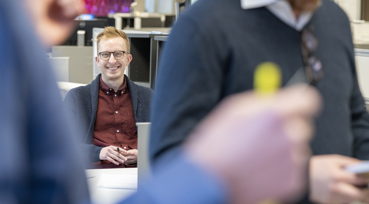 Man sitting at desk smiling at coworkers in foreground Man sitting at desk smiling at coworkers in foreground